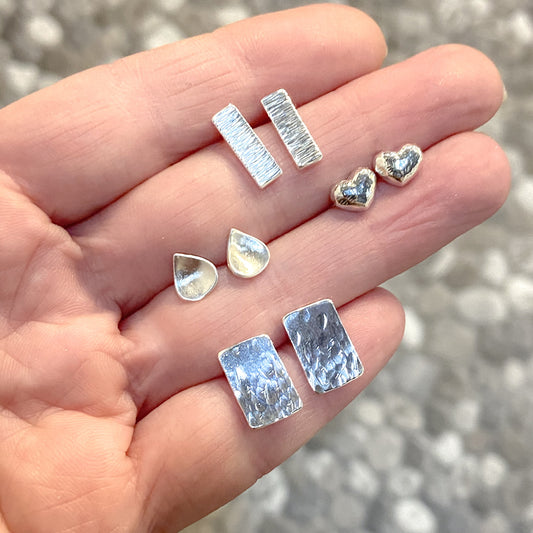 Four different pair of silver stud earring placed between fingers. water drop studs, heart studs, textured bar and rectangle studs.