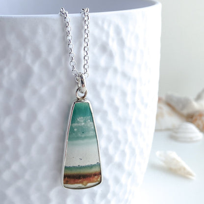 A beautiful opalized wood gemstone pendant that depicts a beach landscape is displayed on a white textured vase with seashells in the background
