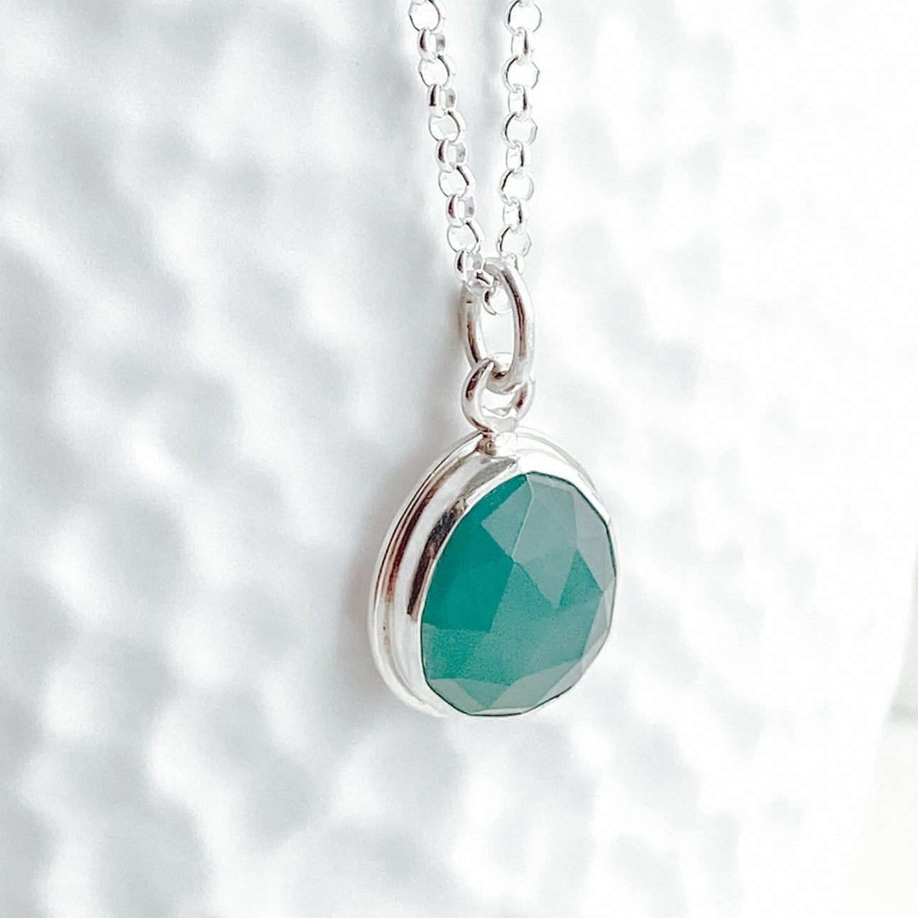 Close up view of a teal green gandiderite gemstone and silver pendant on a white textured background.