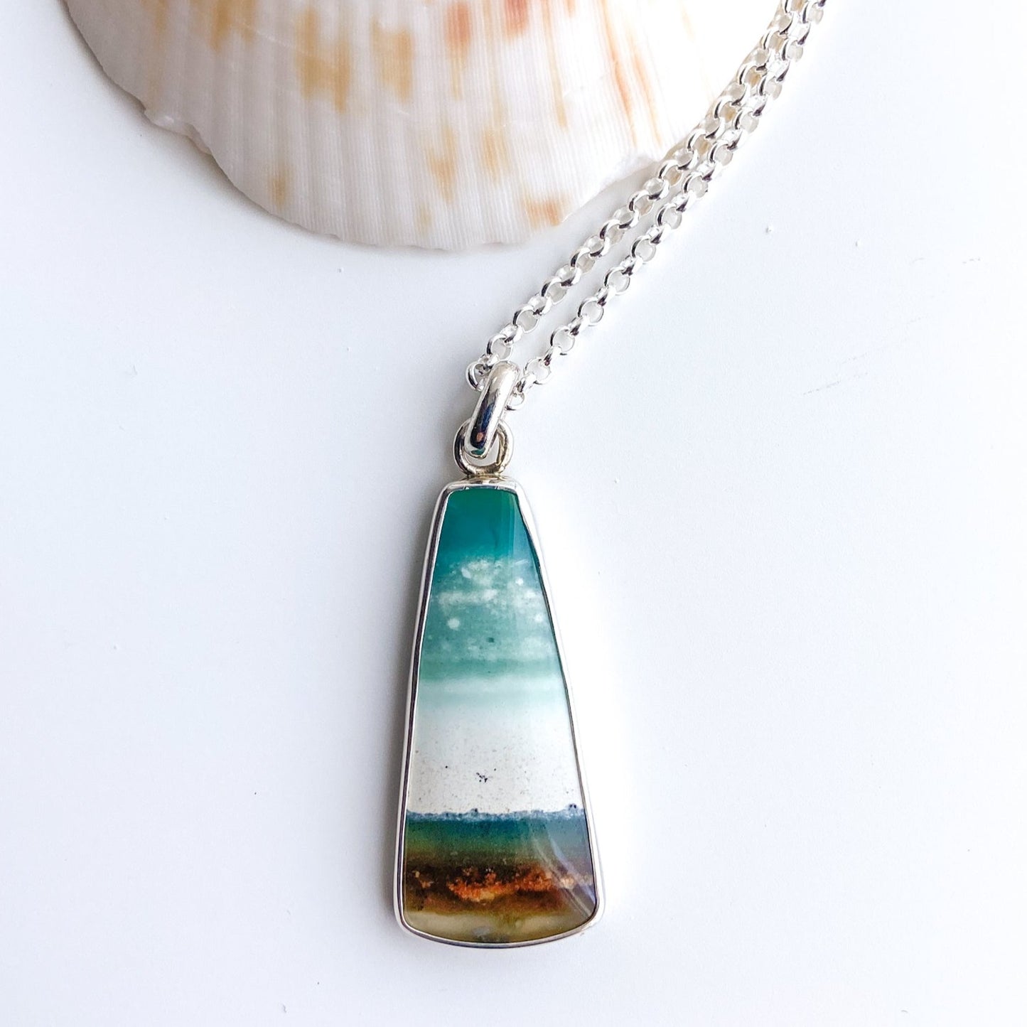 An opalized wood gemstone and silver pendant shows a beach scene in shades of aqua, teal, cream and brown. It is displayed with a scallop shell.