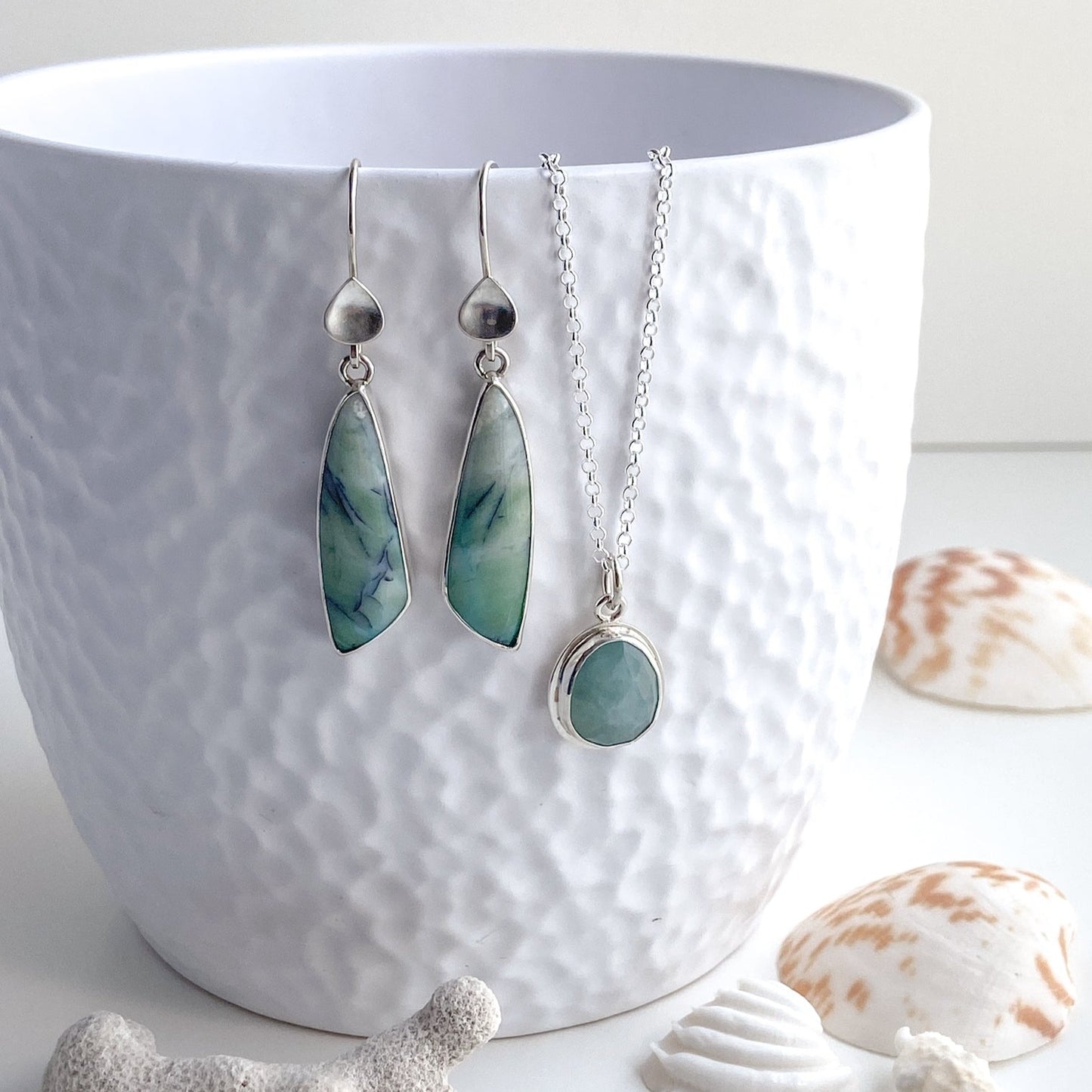 Indonesian opal wood earrings with silver water detail displayed with a sea green Gandiderite pendant on a white vase that is surrounded by seashells