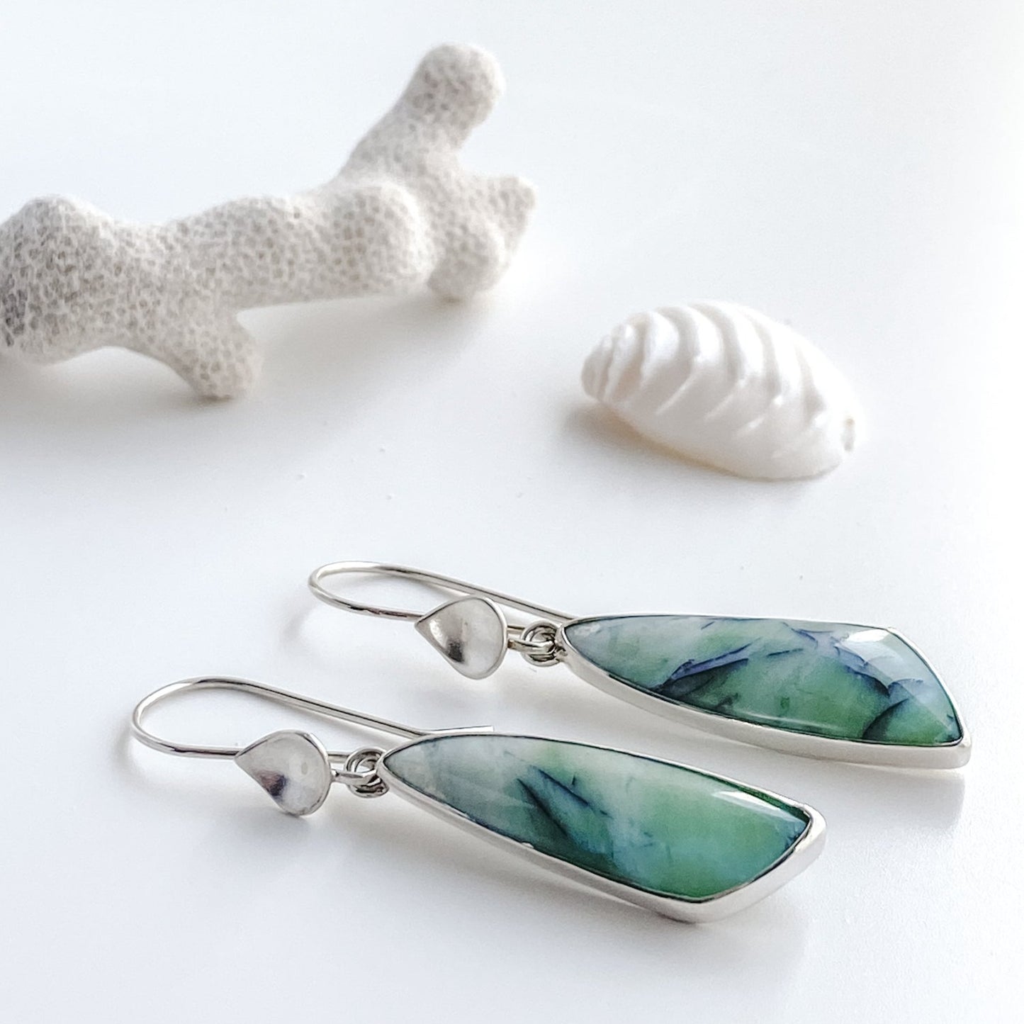 Opalized wood and silver water drop earrings are displayed laying down on a white background with white coral and a white seashell.
