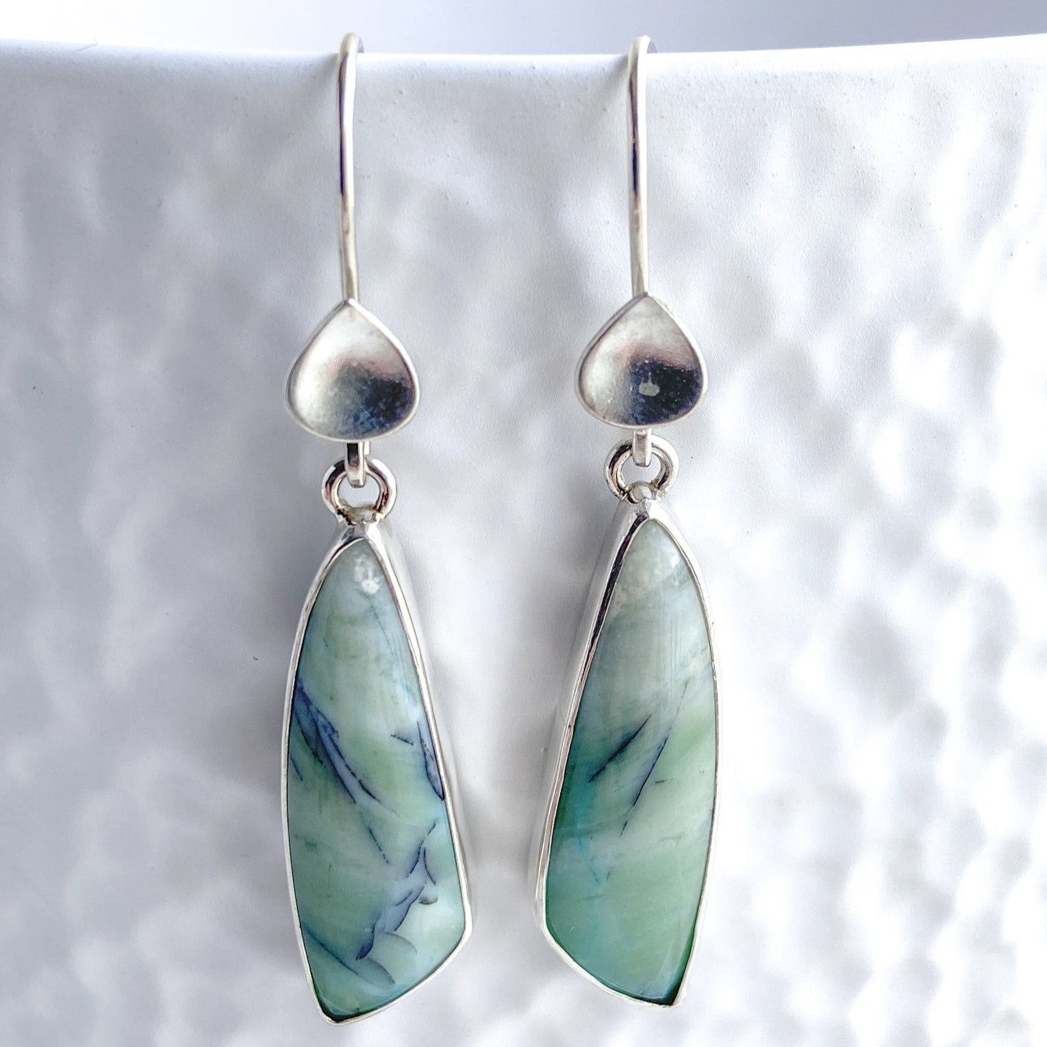 Close-up view of opal wood gemstone earrings with a silver waterdrop embellishment displayed on a white textured background.