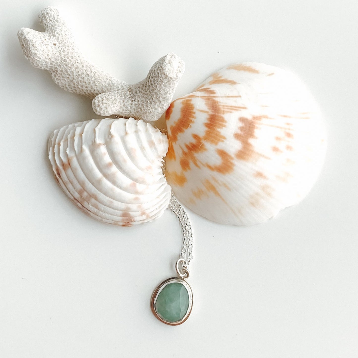Flatlay view of pale green aqua gandiderite  and silver pendant displayed with two seashells and and piece of white coral on a white background.