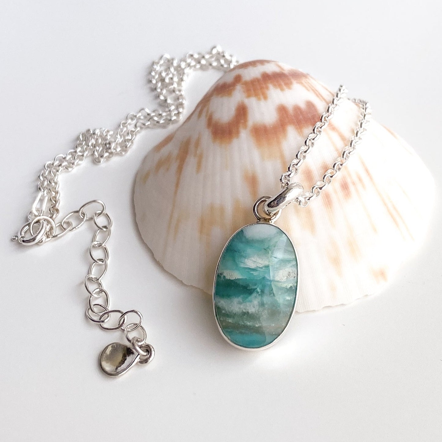 Ocean blue opalized wood and silver pendant draped around a scallop shell on a white background.