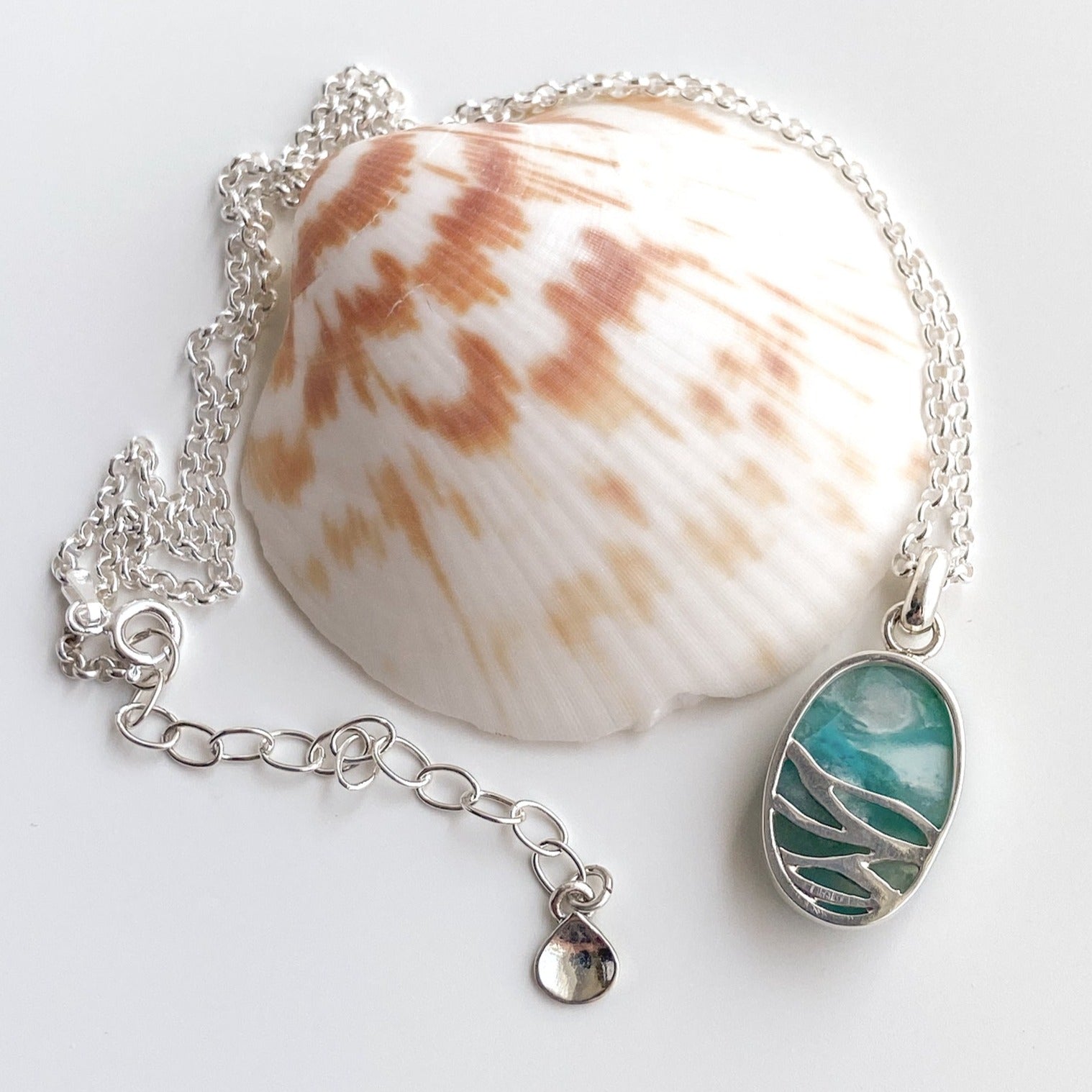 Reverse side view of the ocean blue opalized wood and silver pendant draped around a scallop shell on a white background.